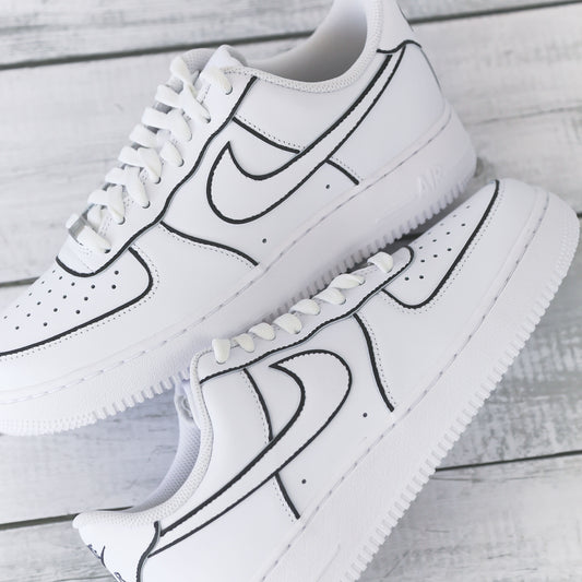 Nike Air Force 1 blanche customisée avec contours noirs brodés, effet dessin, posée sur un fond bois clair