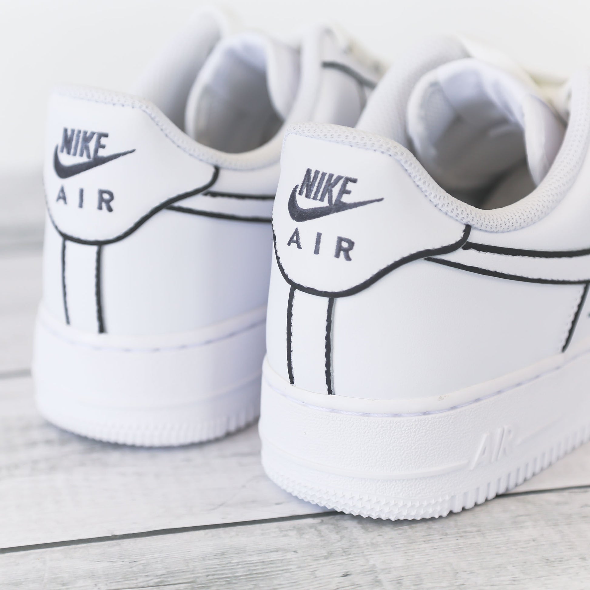 Nike Air Force 1 blanche customisée avec contours noirs brodés, effet dessin, posée sur un fond bois clair